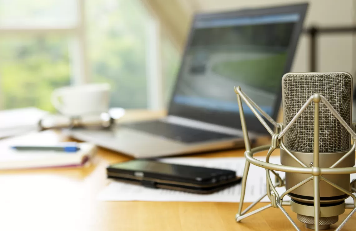 A desk set up with a microphone in the foreground and a laptop, phone, paper and pencil, glasses, and a mug