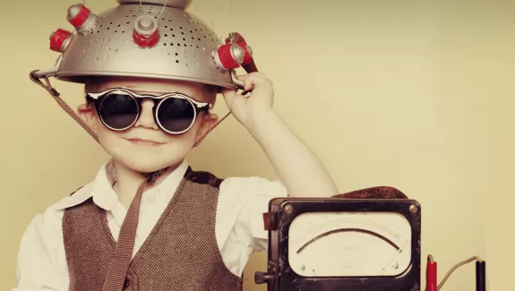 A young boy wearing goggles with a metal colander on his head sitting next to a machine 