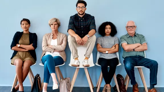 five people sitting in a row of chairs, the third person is sitting on the back of the chair instead of the seat